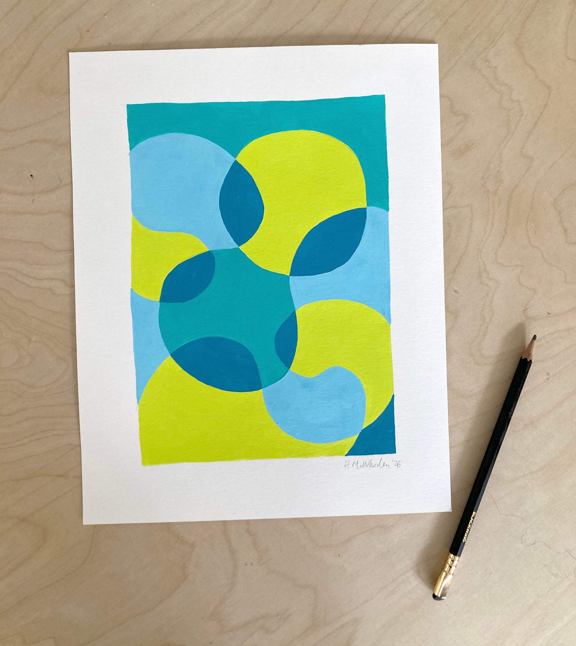 Abstract art print with blue and green shapes on a white background, placed on a wooden surface with a pencil beside it.