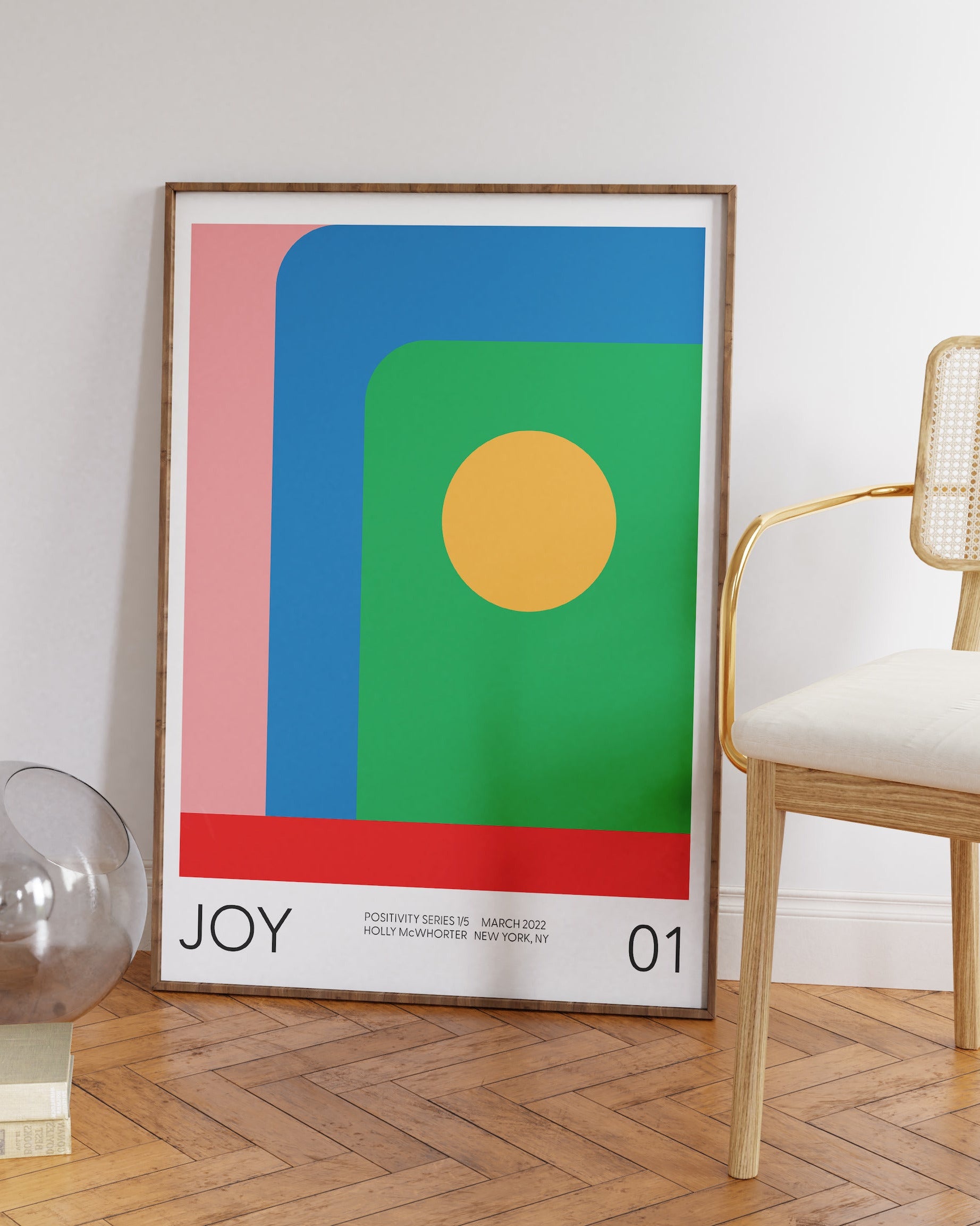 A colorful abstract fine art giclée print with the word 'JOY' featured prominently, displayed above a minimalist wooden chair and a round mirror, against a white wall.